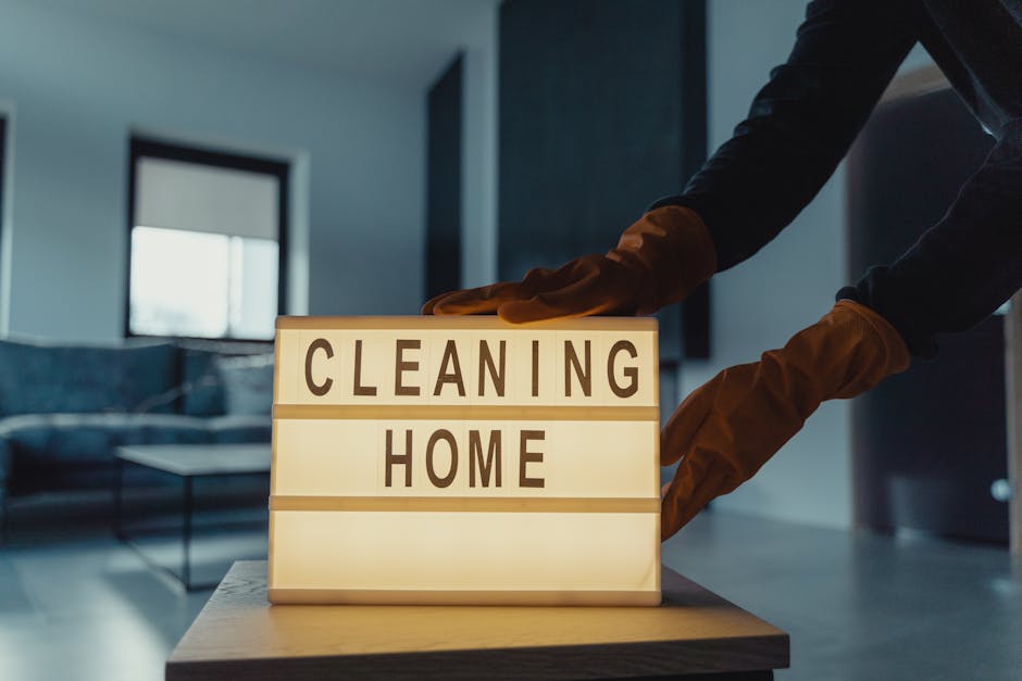 A person wearing brown rubber gloves is adjusting a small illuminated sign on a wooden table that reads 'CLEANING HOME' in a modern, tidy living room. The background features a glass-top coffee table, a dark sofa, and large windows allowing natural light to fill the space, highlighting the clean and polished surfaces. The scene emphasizes domestic surface cleaning and maintenance, reflecting the professional cleaning services offered by Cleaner Kingston for end of tenancy cleaning in KT1, with an emphasis on hygiene and deep cleaning processes.