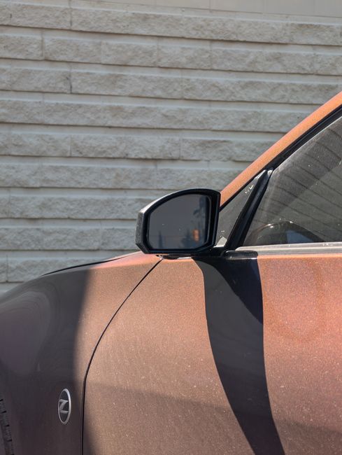 Close-up of the side mirror and front section of a metallic brown car parked near a textured beige brick wall, with sunlight casting shadows on the vehicle. The car's surface appears clean and well-maintained. In the background, the wall's horizontal pattern contrasts with the smooth, reflective surfaces of the car, highlighting the importance of surface cleaning and maintenance. This image exemplifies domestic and commercial cleaning services provided by Cleaner Kingston, focusing on vehicle surface cleanliness as part of comprehensive sanitisation and deep cleaning routines.