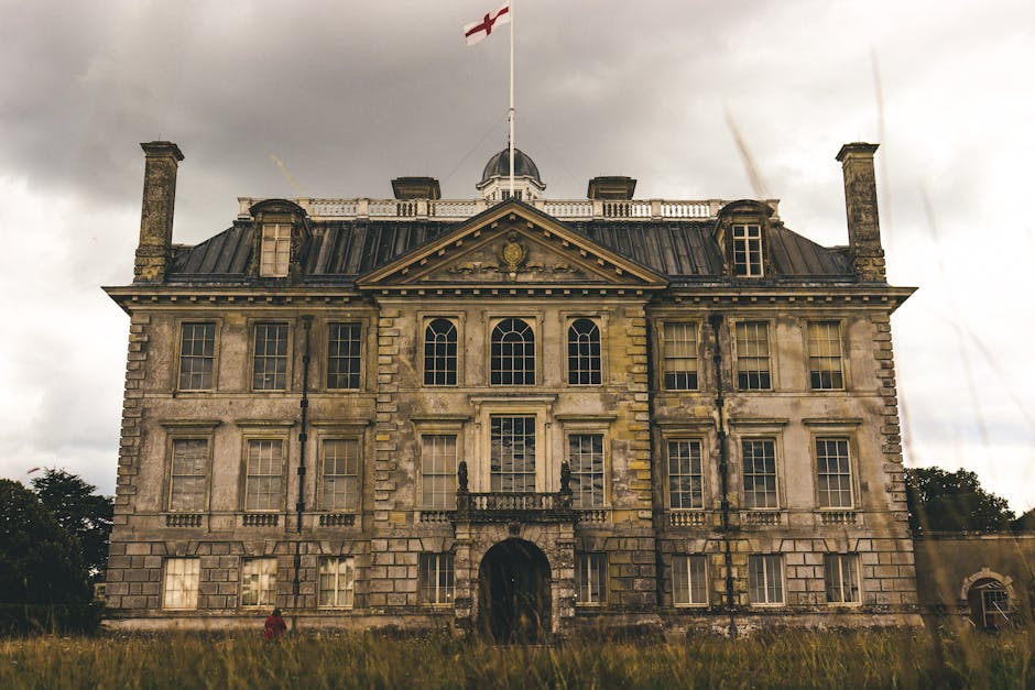 Photograph of an historic, multi-storey stone building with ornate architectural details, tall rectangular windows, and a central arched doorway. The building features a pitched, slate roof with chimneys and a flagpole on top displaying the English flag. A small dome is visible above the roofline. The facade shows weathering and discoloration, with some areas of moss or grime accumulation. The scene is overcast with gray clouds, and the building is surrounded by a grassy area with sparse trees in the background. The image exemplifies an old, grand structure, consistent with architectural preservation and restoration themes, relevant to cleaning and maintenance services highlighted on cleanerkingston.co.uk.