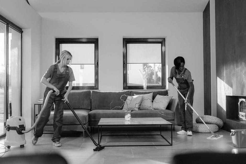 Two professional cleaners from Cleaner Kingston are performing surface cleaning and deep cleaning in a modern living room. They are using vacuum cleaners and mops to clean the wooden floor and the fabric sofa, which is dust-free and well-maintained. The room features two large windows allowing natural light, and the walls are painted in neutral tones with one wall appearing darker for accent. The coffee table in the center has a glass top, and there are cushions on the sofa, indicating a tidy and hygienic environment. The space is bright, clean, and organized, with no visible clutter or dirt, exemplifying effective domestic cleaning practices. The image highlights the importance of proper cleaning tools and methods for maintaining a hygienic living space, aligned with best practices discussed in the page about avoiding common mistakes when booking cleaners in Kingston, as provided by Cleaner Kingston.