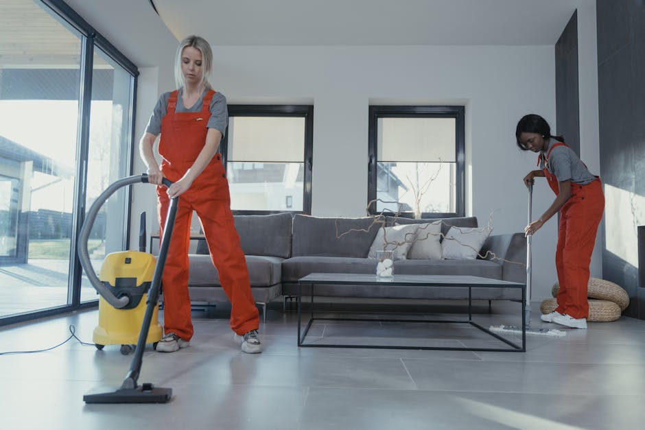 Two professional cleaners from Cleaner Kingston wearing orange overalls are conducting surface cleaning in a modern living room with a grey couch, glass coffee table, and large windows. The cleaner on the left is using a yellow vacuum cleaner to clean the tiled floor, which appears dust-free and shiny. The cleaner on the right is using a mop to wipe the floor near a pouf, with some cleaning cloths and accessories in the background. The room is well-lit with natural light from the windows, highlighting the clean and tidy appearance of the space. This image exemplifies effective deep cleaning and sanitisation practices suited for domestic cleaning environments, ensuring a hygienic and well-maintained living space, aligning with the tips discussed in the page 'Common mistakes when booking cleaners in Kingston what to avoid, KINGSTON' on cleanerkingston.co.uk.