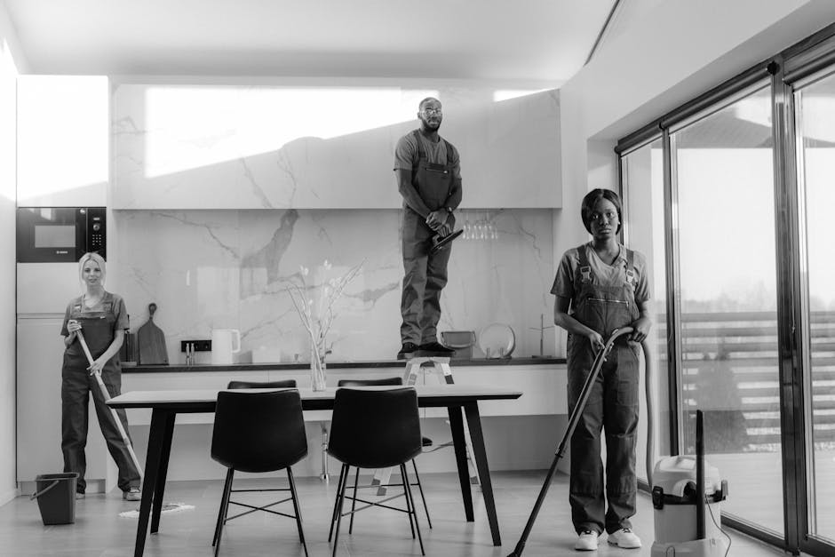 Two professional cleaners from Cleaner Kingston are performing surface cleaning and deep cleaning in a modern living room. They are using vacuum cleaners and mops to clean the wooden floor and the fabric sofa, which is dust-free and well-maintained. The room features two large windows allowing natural light, and the walls are painted in neutral tones with one wall appearing darker for accent. The coffee table in the center has a glass top, and there are cushions on the sofa, indicating a tidy and hygienic environment. The space is bright, clean, and organized, with no visible clutter or dirt, exemplifying effective domestic cleaning practices. The image highlights the importance of proper cleaning tools and methods for maintaining a hygienic living space, aligned with best practices discussed in the page about avoiding common mistakes when booking cleaners in Kingston, as provided by Cleaner Kingston.