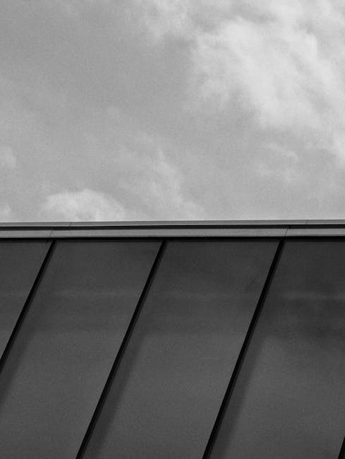 Close-up view of a modern building's exterior featuring large, reflective glass panels arranged in a diagonal pattern, with dark metal framing. The surface of the glass is clean and free of smudges, providing a clear reflection of the overcast sky above. The lighting highlights the smooth texture and sleek finish of the panels, emphasizing the meticulous surface cleaning and maintenance typical of professional sanitisation and deep cleaning services. This image captures the emphasis on surfaces and materials like glass and metal, aligned with Kingston end of tenancy cleaning practices, as promoted by Cleaner Kingston in their professional cleaning and hygiene solutions.