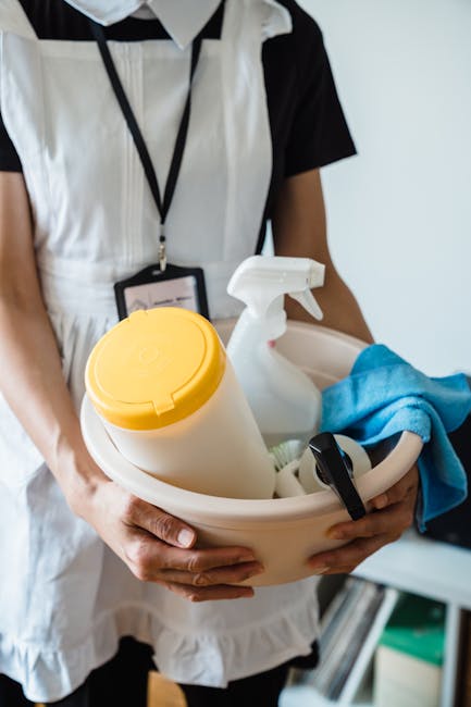 A person wearing brown cleaning gloves is adjusting a small illuminated sign on a wooden table that reads 'CLEANING HOME' in black uppercase letters. The background shows a modern living room with a sofa, a coffee table, and large windows allowing natural light to fill the space. The room appears clean and tidy, emphasizing surface cleaning and maintenance. This scene relates to domestic cleaning services offered by Cleaner Kingston, highlighting professional cleaning practices suitable for end of tenancy or deep cleaning to ensure hygienic living environments.