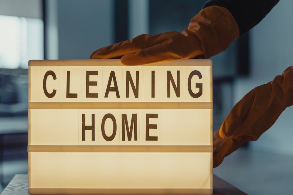 A person wearing brown cleaning gloves is placing a lightbox sign on a white surface that displays the words 'CLEANING HOME' in brown block letters, indicating domestic cleaning services. The backgrou
