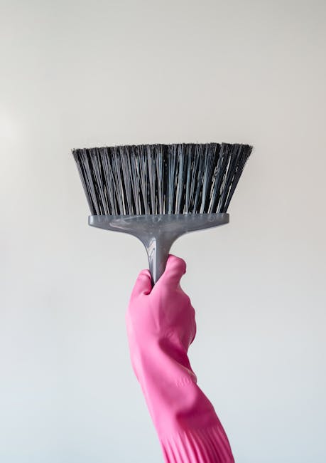 A person wearing a pink rubber glove is holding a black plastic dustpan with rigid bristles, positioned against a plain white wall. The surface appears clean with no visible dust or debris, suggesting recent surface cleaning or preparation for dusting. The lighting is bright and neutral, highlighting the smooth, matte texture of the glove and the clean surfaces of the dustpan. This image emphasizes the cleaning tools used in surface cleaning, part of domestic cleaning services offered by Cleaner Kingston near Kingston Bridge, focusing on hygiene and maintenance in a residential setting.