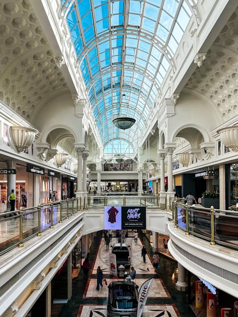 An indoor shopping mall featuring a large, curved glass ceiling that allows natural light to illuminate the space. The ceiling is supported by white ornate arches and columns, with decorative floral details along the edges. On the upper level, polished wooden handrails run along the balcony overlooking the ground floor, where glossy tiled flooring displays a patterned design. Storefronts with illuminated signs line the mall, and shoppers are walking on both levels. The overall environment appears clean, bright, and well-maintained, reflecting professional cleaning standards promoted by Cleaner Kingston for commercial spaces like shopping centres.
