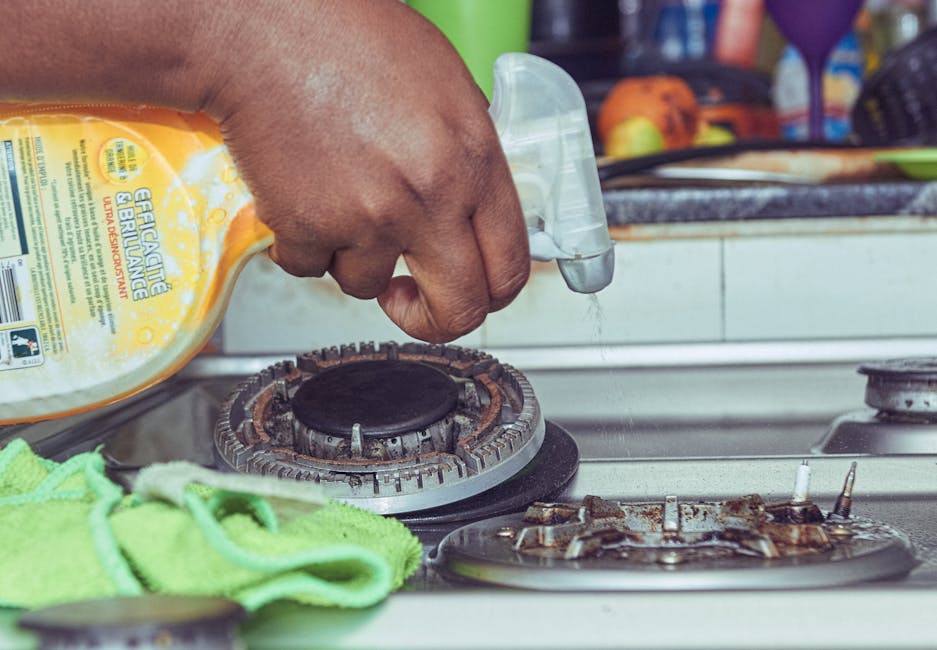 A close-up view of a person's hand holding a spray bottle, applying a cleaning solution to the stovetop of a gas hob in a domestic kitchen. The stovetop has visible signs of grease and grime, with burnt residues on the burners. The kitchen counter area includes a yellow cleaning product bottle with a trigger spray, a green cloth, and other cleaning supplies nearby. The background features a tiled backsplash, with some kitchen utensils and colorful items slightly out of focus. The image emphasizes surface cleaning and deep kitchen sanitisation processes conducted by Cleaner Kingston, highlighting effective domestic cleaning efforts for kitchens on Clarence Street, Kingston.