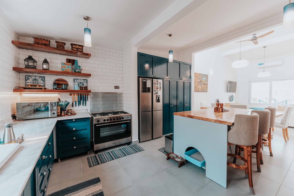 A bright, modern kitchen featuring a stainless steel refrigerator, a gas stove, and blue cabinetry beneath a light-colored countertop. Open wooden shelves mounted on a white brick backsplash hold decorative items, jars, and a microwave oven. The countertop area is clean and free of clutter, with a dishcloth hanging nearby and a small woven rug in front of the stove. An island with a wooden top extends into the dining area, surrounded by beige upholstered chairs. The room is well-lit with natural light pouring through large windows, and there are pendant ceiling lights providing additional illumination. The overall scene depicts a hygienic, well-maintained kitchen surface suitable for deep cleaning and surface sanitisation, representative of services offered by Cleaner Kingston.