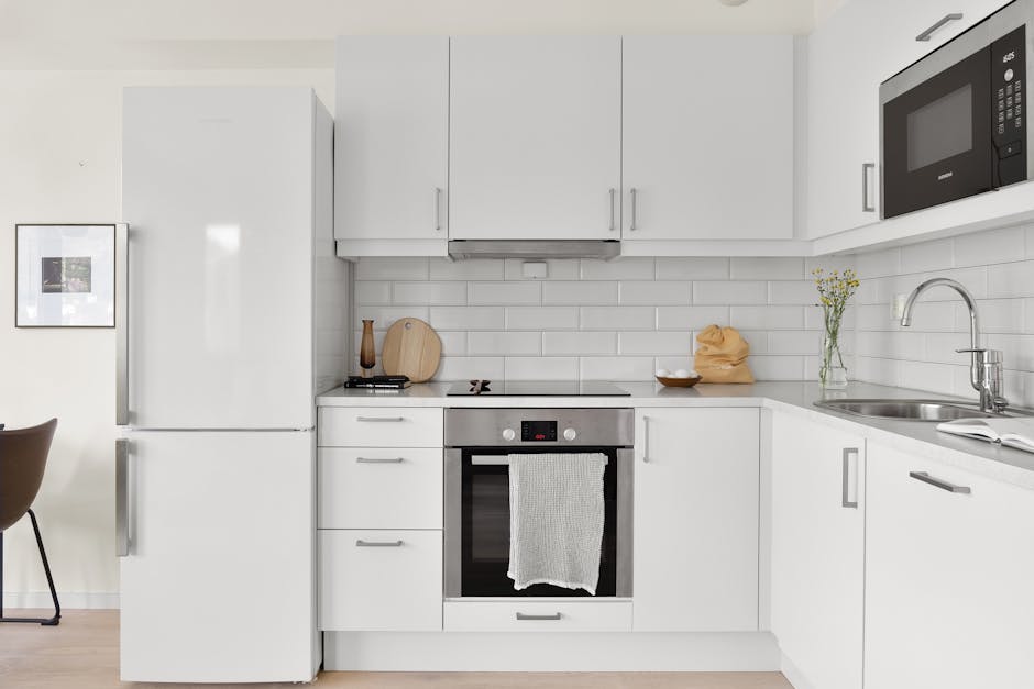 A modern, tidy kitchen with white cabinetry, a stainless steel oven and microwave, and a white tiled backsplash. The countertops are clear, with a few decorative items including a wooden cutting board, a small bowl, and a vase with flowers. The refrigerator is white and situated on the left side of the image. The lighting highlights the clean and polished surfaces, with no visible dirt or clutter, illustrating a high standard of surface cleaning and sanitisation provided by Cleaner Kingston. The arrangement and materials emphasize a hygienic and well-maintained domestic space.