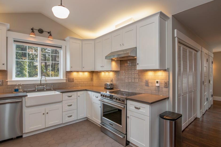 A bright, modern kitchen featuring a stainless steel refrigerator, a gas stove, and blue cabinetry beneath a light-colored countertop. Open wooden shelves mounted on a white brick backsplash hold decorative items, jars, and a microwave oven. The countertop area is clean and free of clutter, with a dishcloth hanging nearby and a small woven rug in front of the stove. An island with a wooden top extends into the dining area, surrounded by beige upholstered chairs. The room is well-lit with natural light pouring through large windows, and there are pendant ceiling lights providing additional illumination. The overall scene depicts a hygienic, well-maintained kitchen surface suitable for deep cleaning and surface sanitisation, representative of services offered by Cleaner Kingston.
